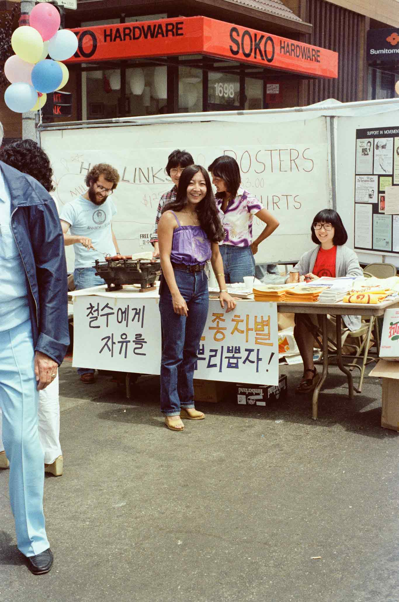 Individuals gather around a table cooking hot dogs and handing out t-shirts. Posters with Korean characters written on them are hung on the table.
