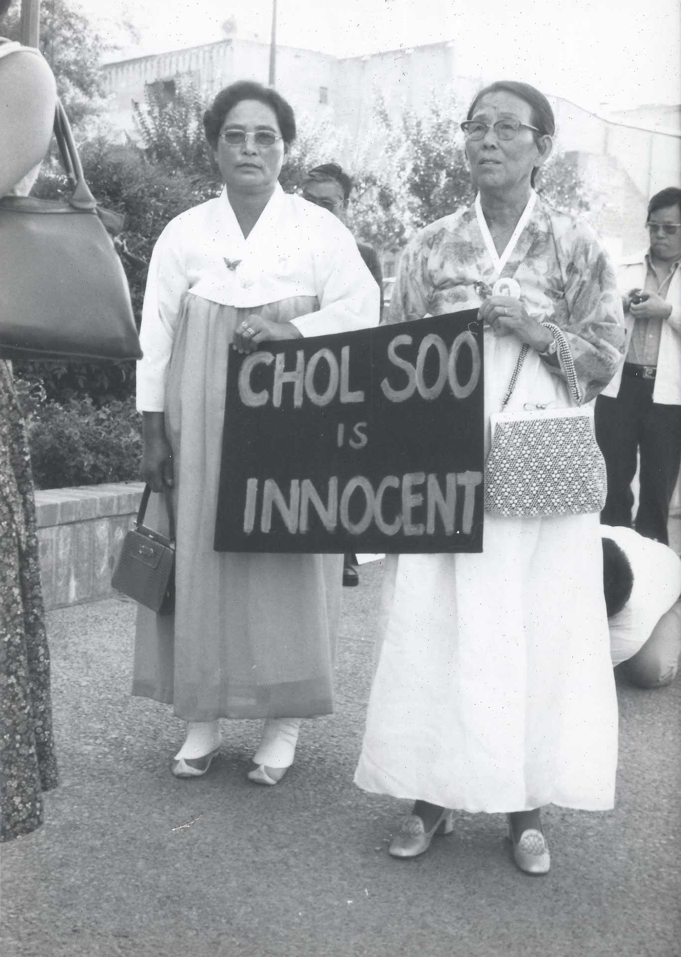 Two women stand side by side outdoors, each wearing traditional Korean hanbok dresses and holding handbags. They are holding a large sign that reads "CHOL SOO IS INNOCENT." Other people are partially visible in the background, and the setting appears to be a public demonstration or gathering.
