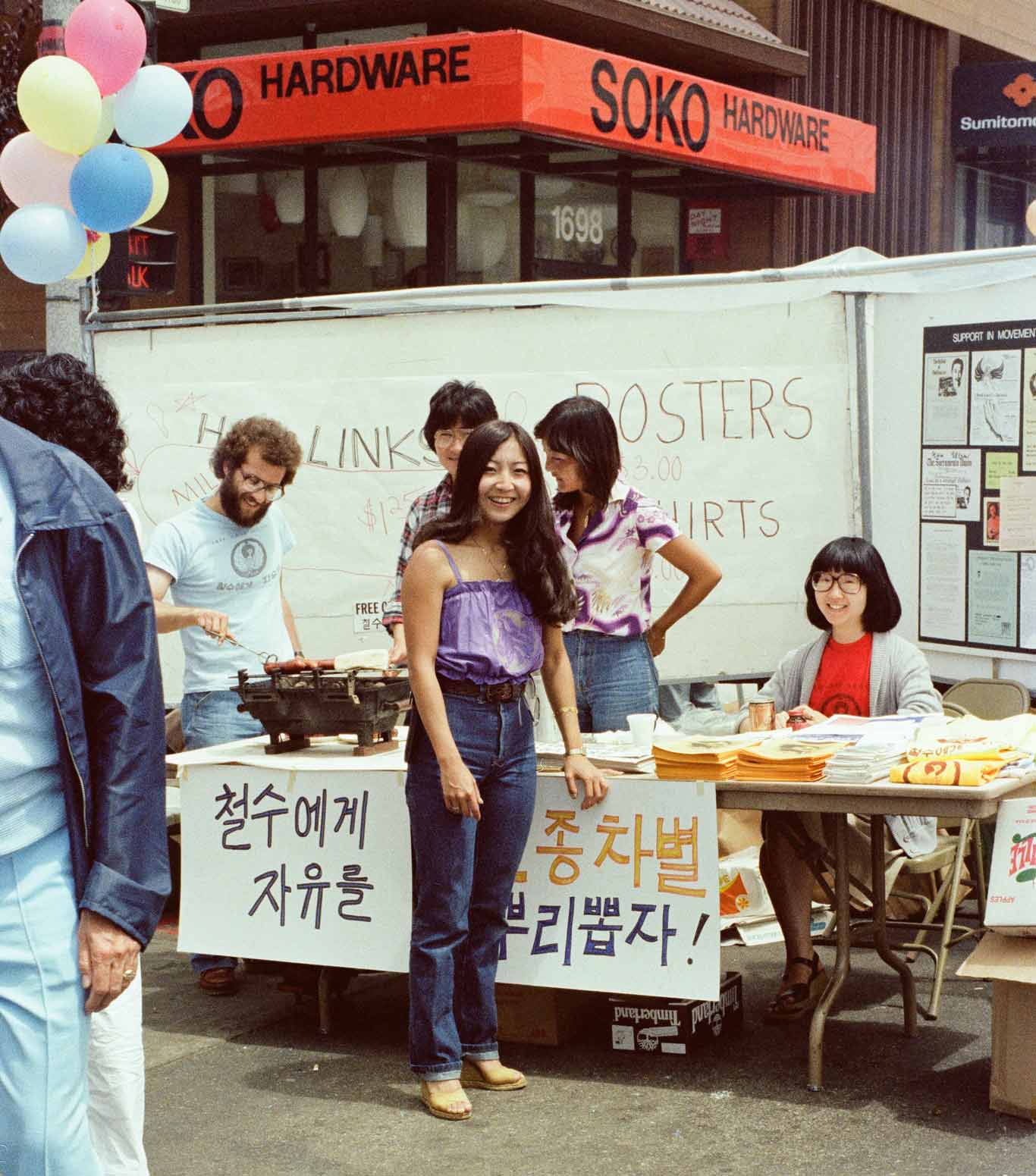 Individuals gather around a table cooking hot dogs and handing out t-shirts. Posters with Korean characters written on them are hung on the table.
