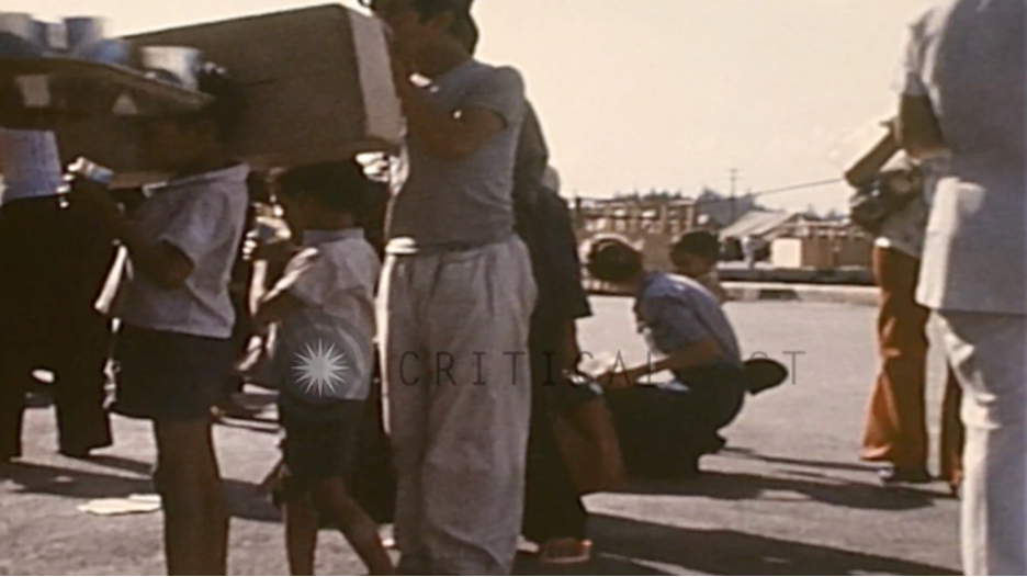 Operation New Life in the United States. Vietnamese refugees carrying their personal belongings wait in a line for processing. Civilians and navy personnel serve refreshments to the refugees waiting in the line. Vietnamese men, women and children walk wearing shower slippers provided by U.S. personnel.