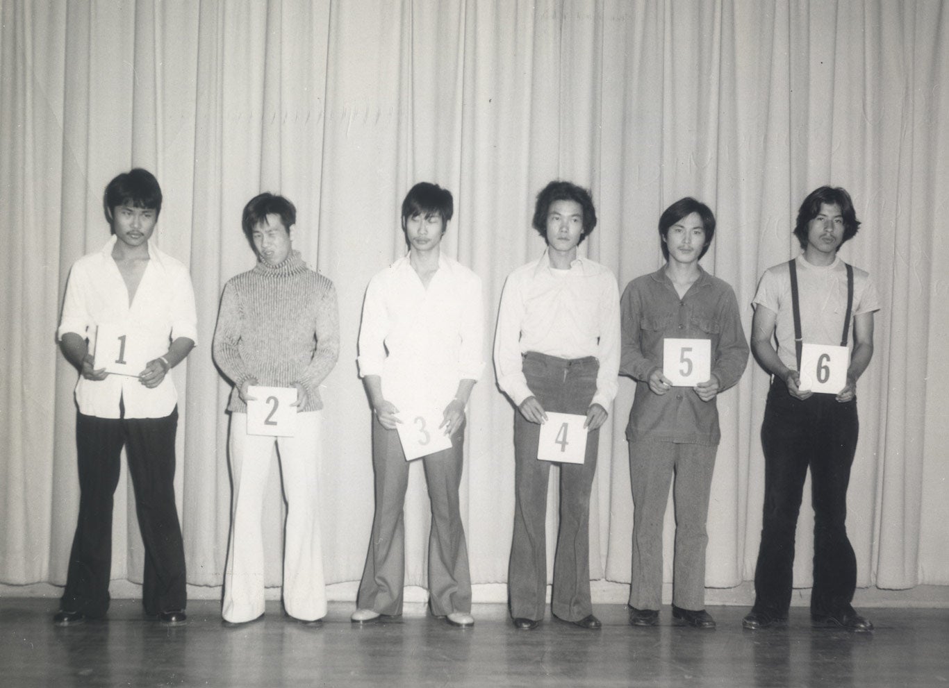 Six men stand in a line on a stage in front of a curtain, each holding a card with a number, starting with 1 on the left and going through to 6 on the right. Suspect number 5 is Chol Soo Lee.