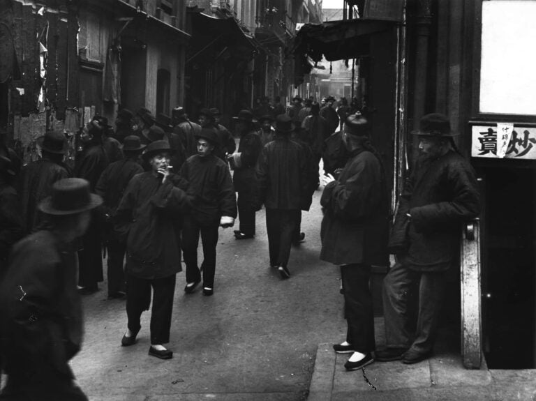 Street life of Ross Alley in San Francisco's Chinatown depicts Chinese men with queue hairstyle, wearing hats, and shanku - shirts and trousers.