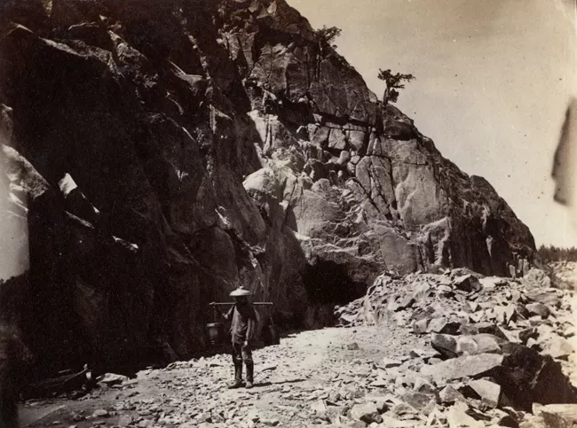 A Chinese man holds a carrying pole with two buckets on either end over his shoulders standing between a mountainside and piles of rubble.