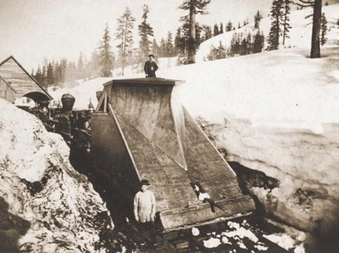 A man stands atop a snow plow affixed to the front of a train while a Chinese worker stands at the bottom as it paves a path through deep snow.