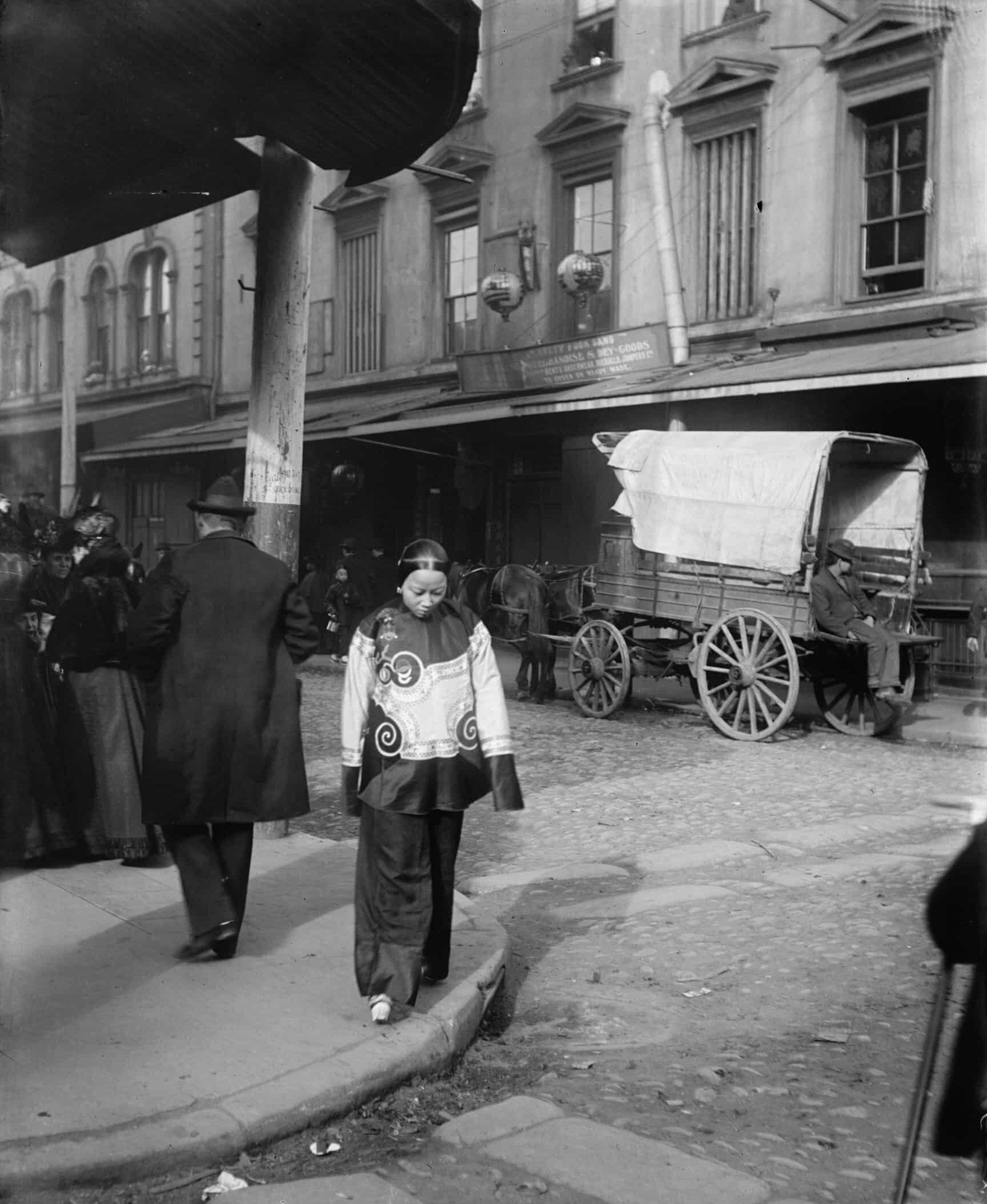 Chinese woman in holiday shanku with chanzu bound feet walking away from a group of white people in Chinatown. Horse-drawn wagon parked on the street.