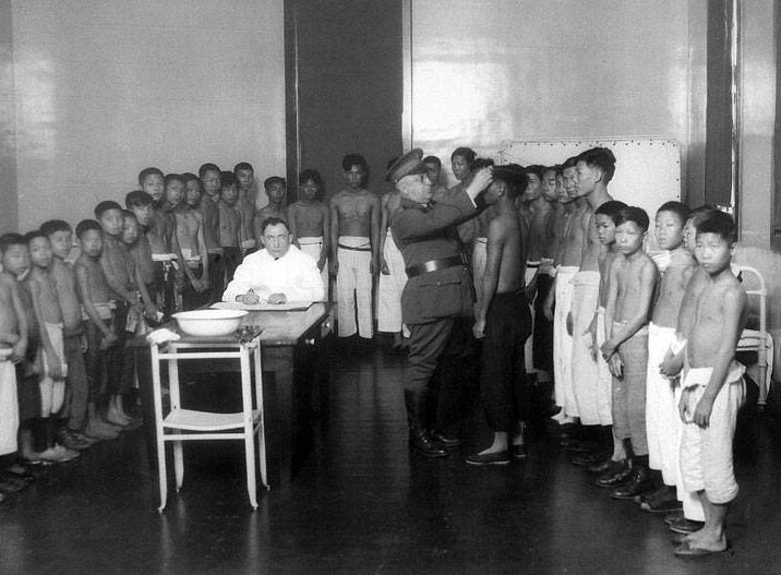 A U.S. Marine examines a Chinese boy's head while a man in a lab coat writes notes. A group of shirtless Chinese boys and young men wait their turn.