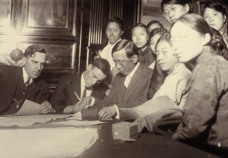 Two white men and a Chinese man in suits are seated at the table processing immigration papers as a group of Chinese women crowd around them.
