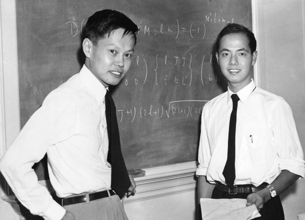 Chinese-born, Nobel Prize recipients Chen Ning Yang on the right and Tsung-Dao Lee on the left stand next to a chalkboard and pose for a photo.