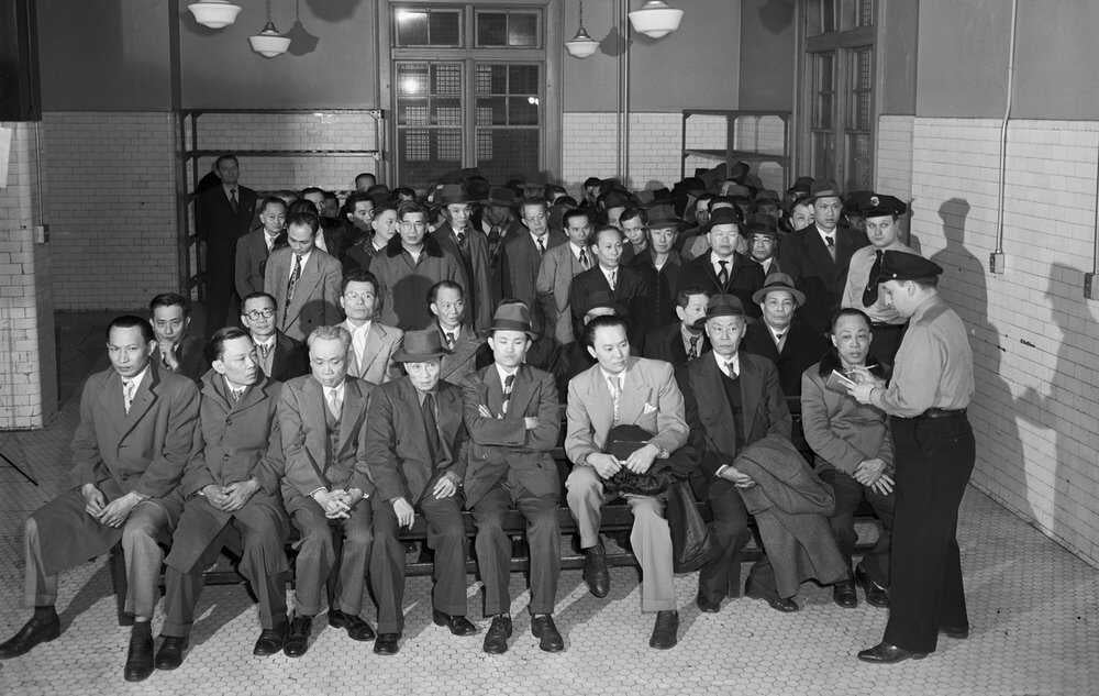 Two white immigration officers interrogate a group of well-dressed Chinese immigrants in a room on Ellis Island.