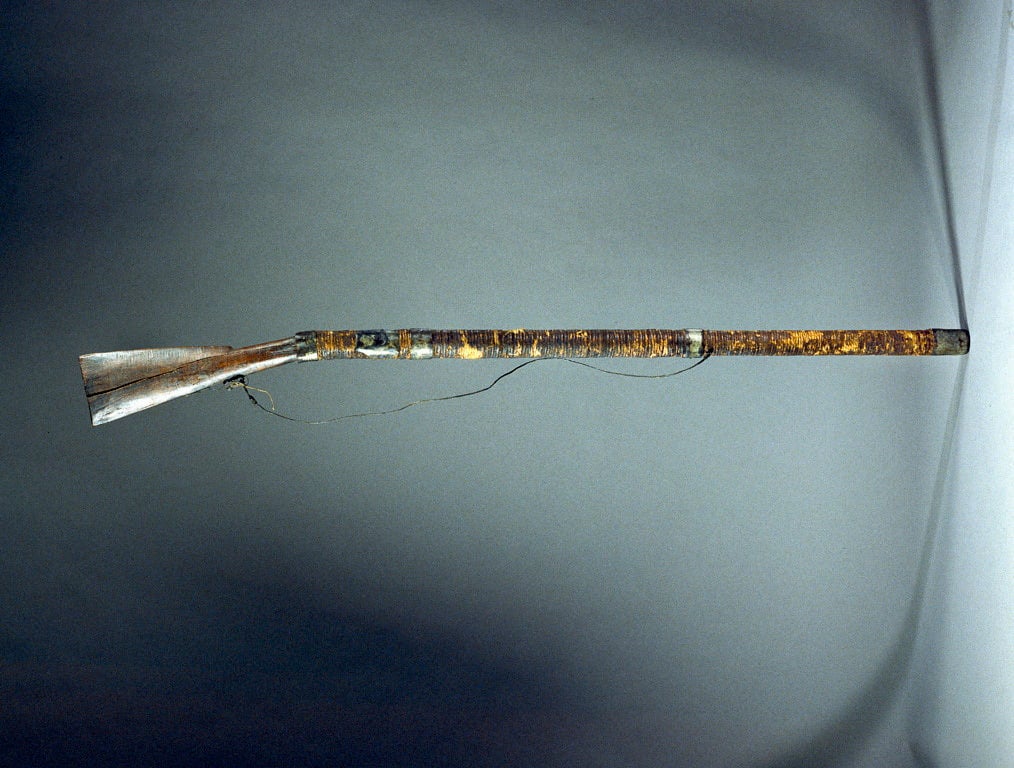 Exhibit photo of a homemade gun from the Philippines. Wood carved handle with a long, narrow barrel and attached strap.