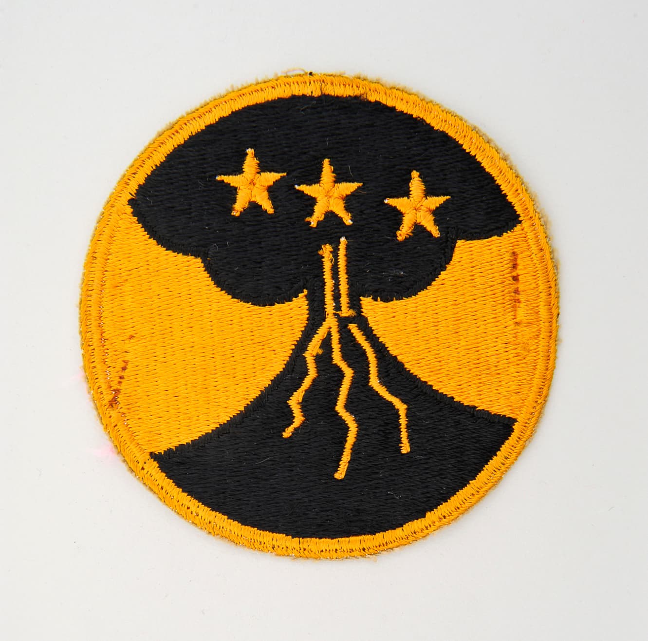 Round military patch depicts a black volcano and a cloud of smoke with 3 yellow stars above against a yellow background.