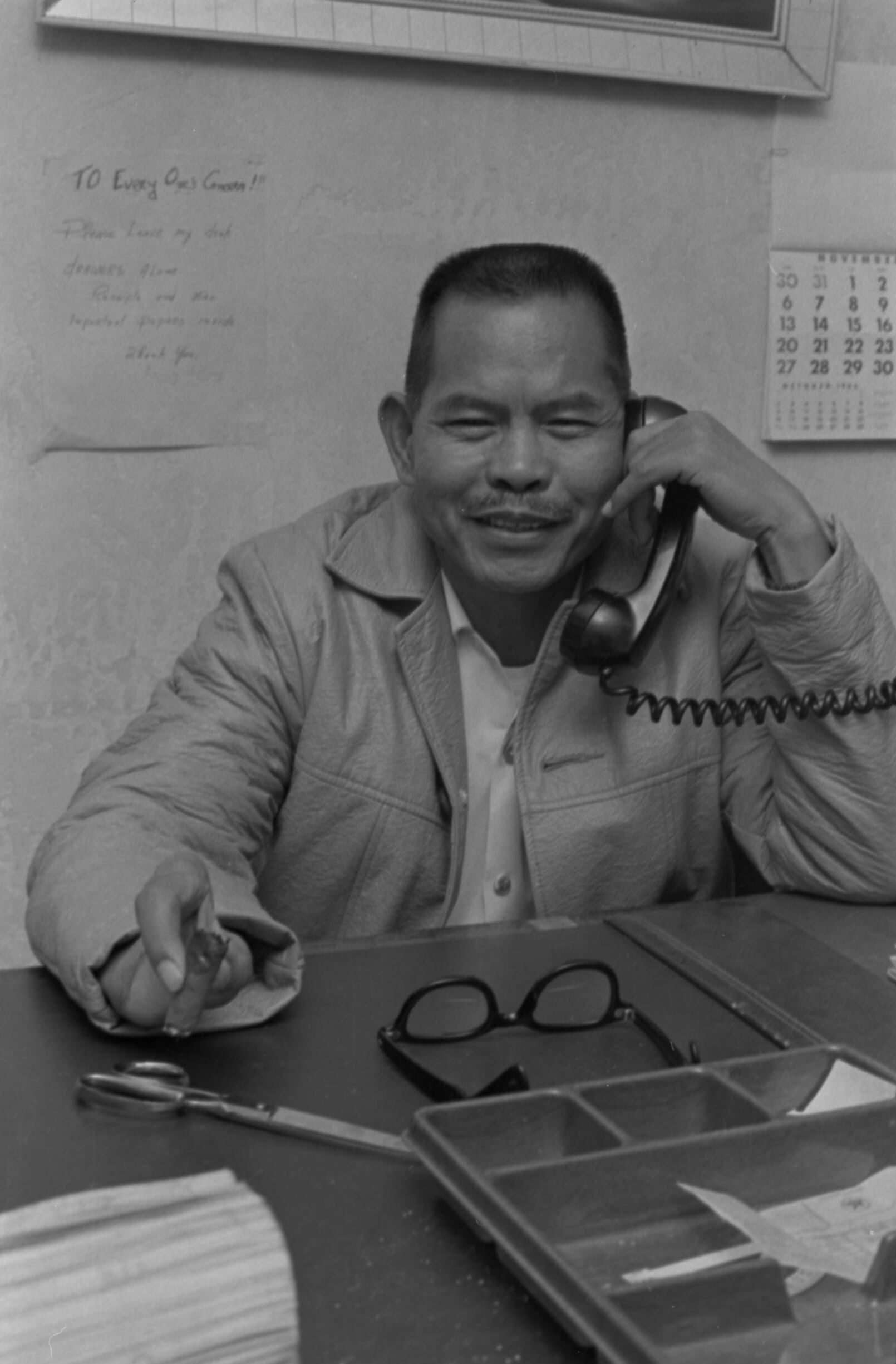 Itliong sitting at his desk, smiling at the camera with the phone to his ear and his signature cigar in the remaining two fingers on his right hand.