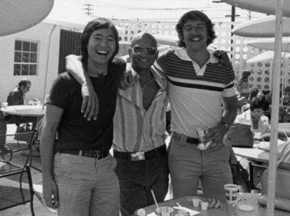Navroze Mody, an Indian American man, wearing sunglasses and an open button down dress shirt, is smiling and standing between two unidentified men.