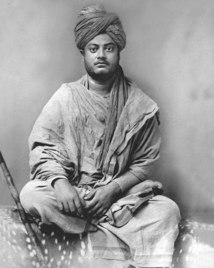 An Indian Hindu Monk Swami Vivekananda dressed in robes and a turban, sits with his right leg crossed over his left thigh.