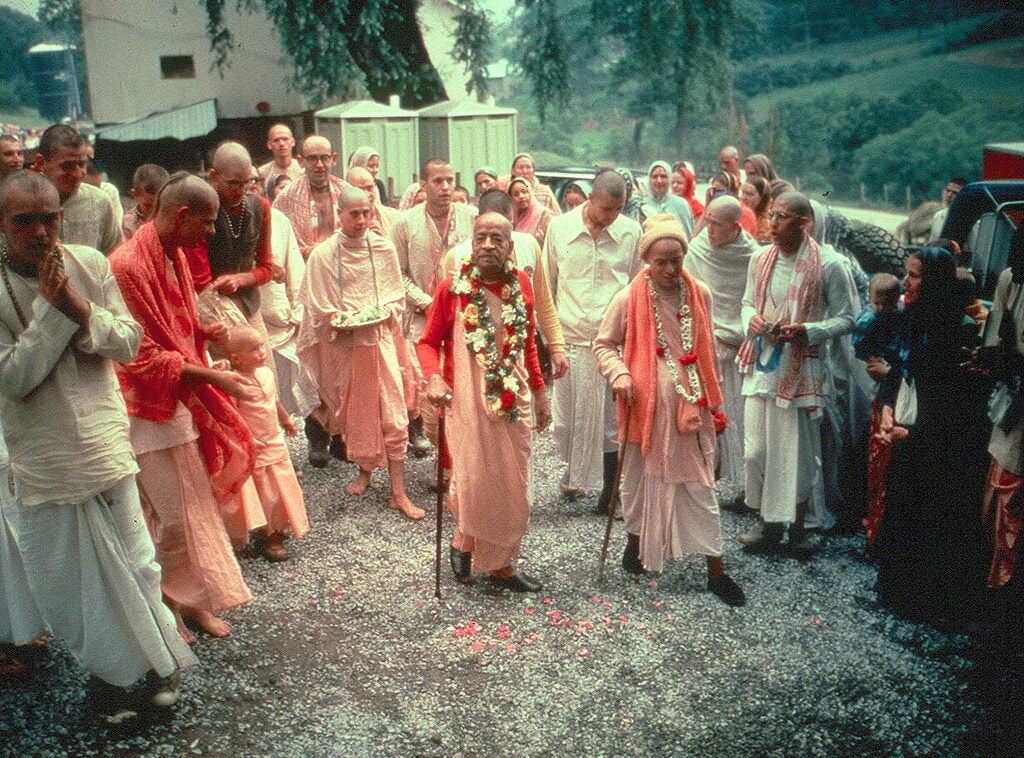 Srila Prabhupada, a spiritual teacher from India, dressed in a robe and flower garlands, walks with Hare Krishna devotees.