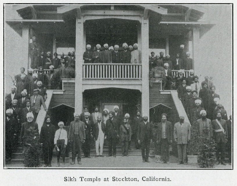 Sikh men wearing turbans, hats, and suits standing along the exterior of the Stockton Gurdwara, a place of Sikh worship and community center.
