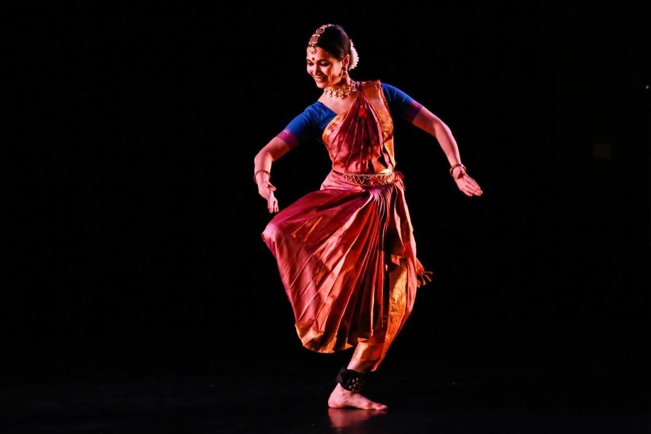 Bharatanatyam dancer in a dance pose in front of a black backdrop wearing an elaborate saree, fitted blouse, and Oddiyanam belt.