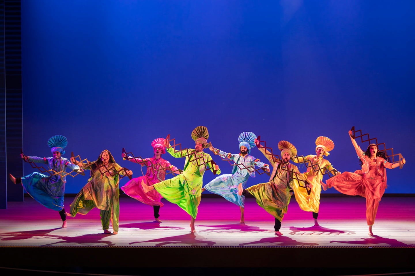 Bhangra dancers in colorful outfits are standing on one leg during a performance on stage while using sapp, a Punjabi musical instrument.