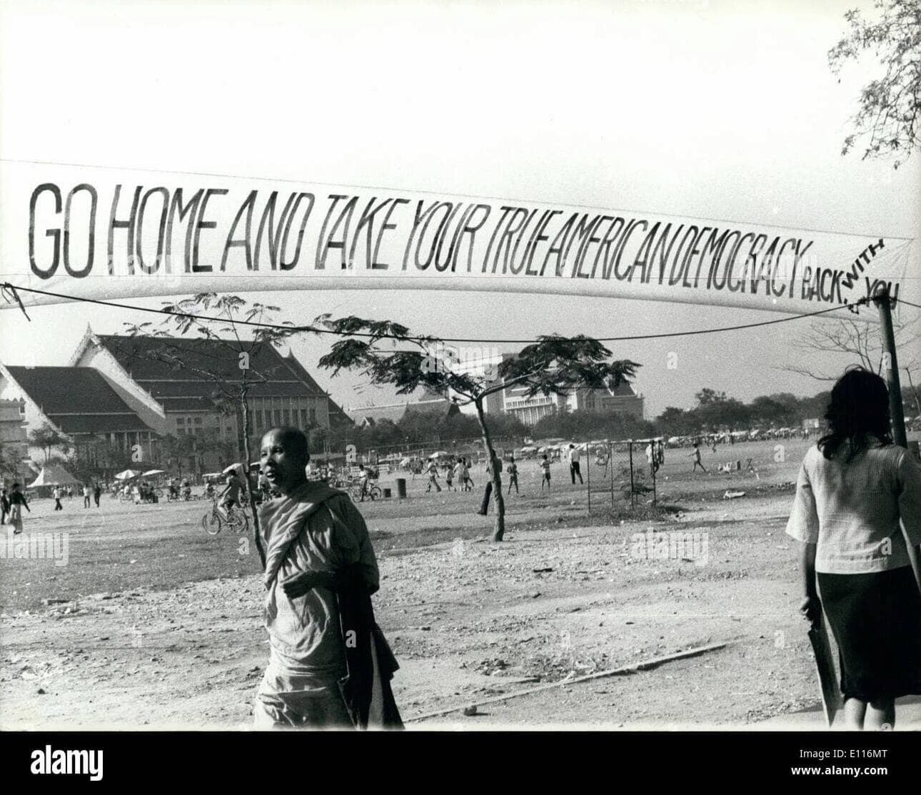 Large banner with text "Go Home and Take Your True American Democracy Back With You" hangs in front of large field crowded with people.