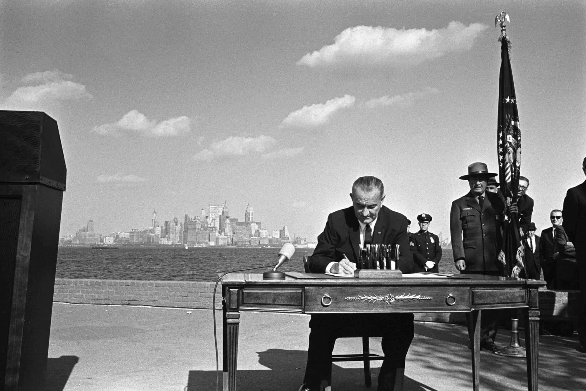 President Lyndon B. Johnson, seated outdoors at desk, signs Immigration Act. Uniformed men and men in suits crowd behind him to the right.