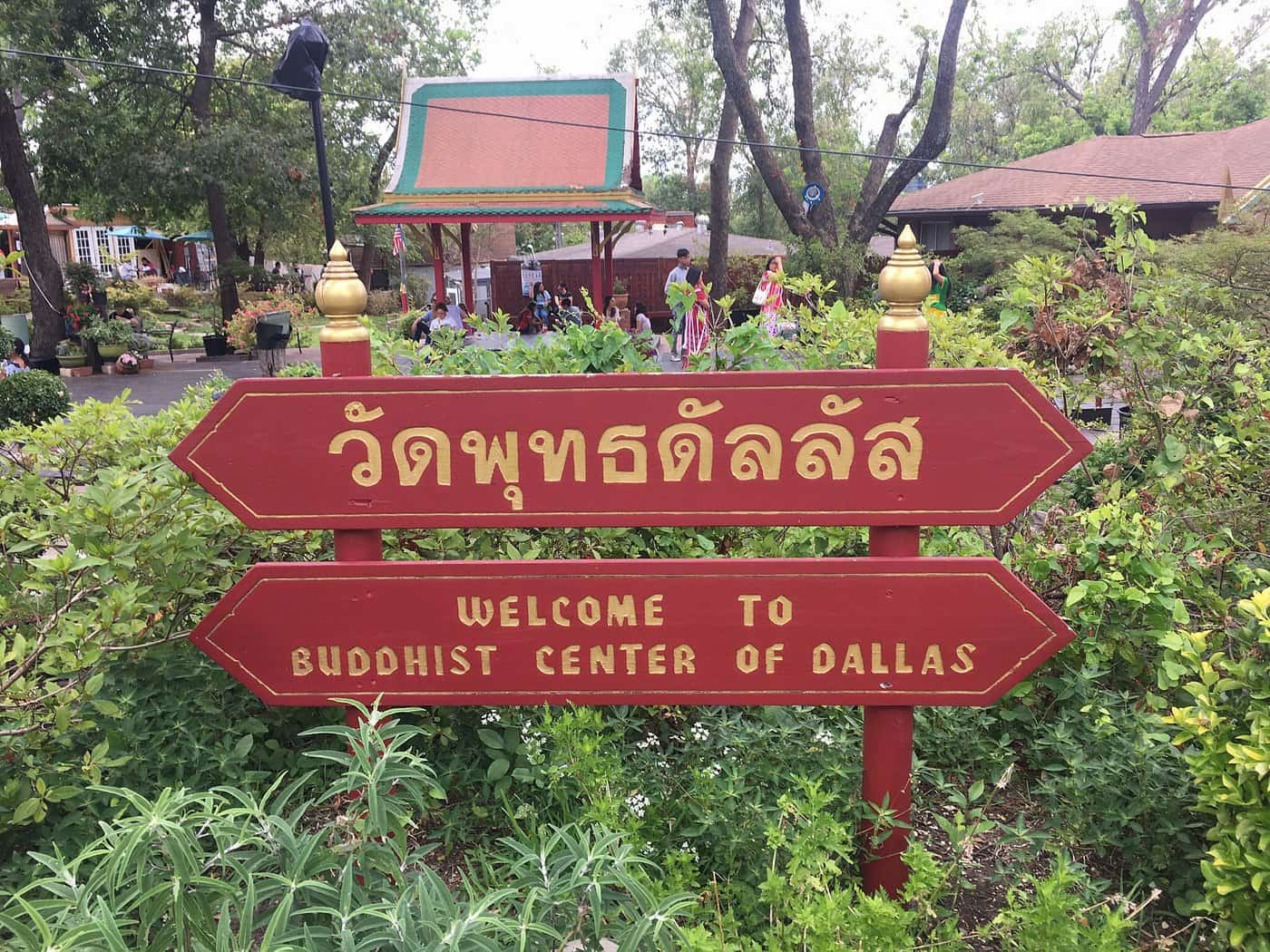 Gold-colored painted words on red wooden signs read "Welcome to Buddhist Center of Dallas" in English and "Dallas Buddhist Center" in Thai.