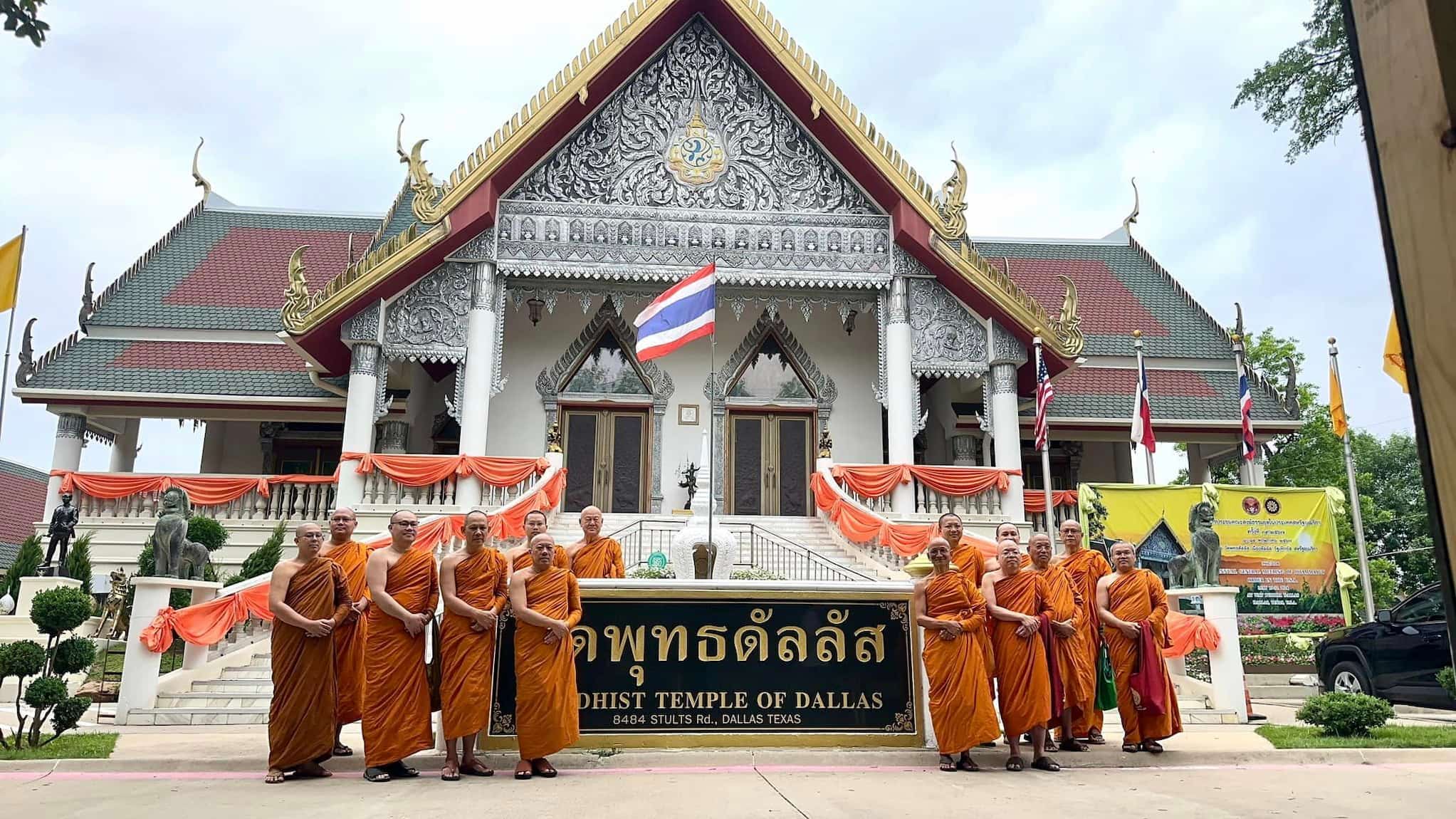 Buddhist monks, dressed in orange robes, stand beside "Buddhist Temple of Dallas" sign. The monks stand in front of steps leading to Temple.