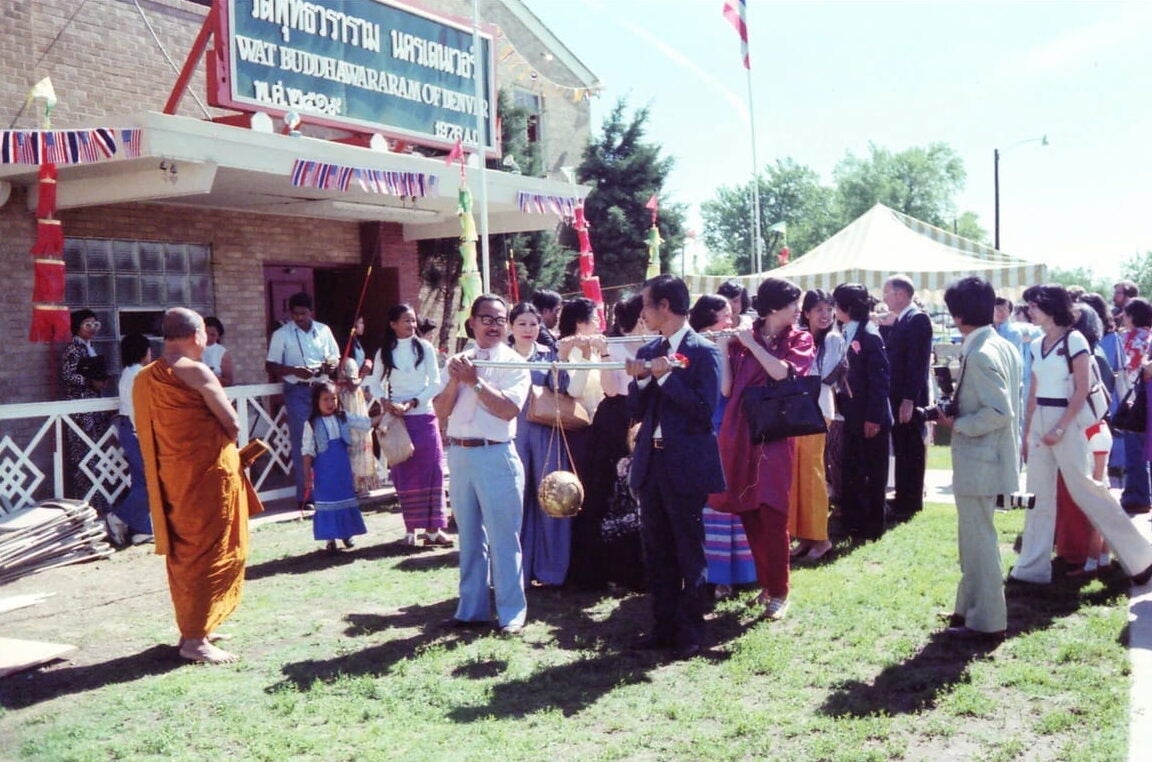 A group of Thai people gather outside a wat. The group stands in a line formation and carries a sphere that is hung from a rod as a monk watches.