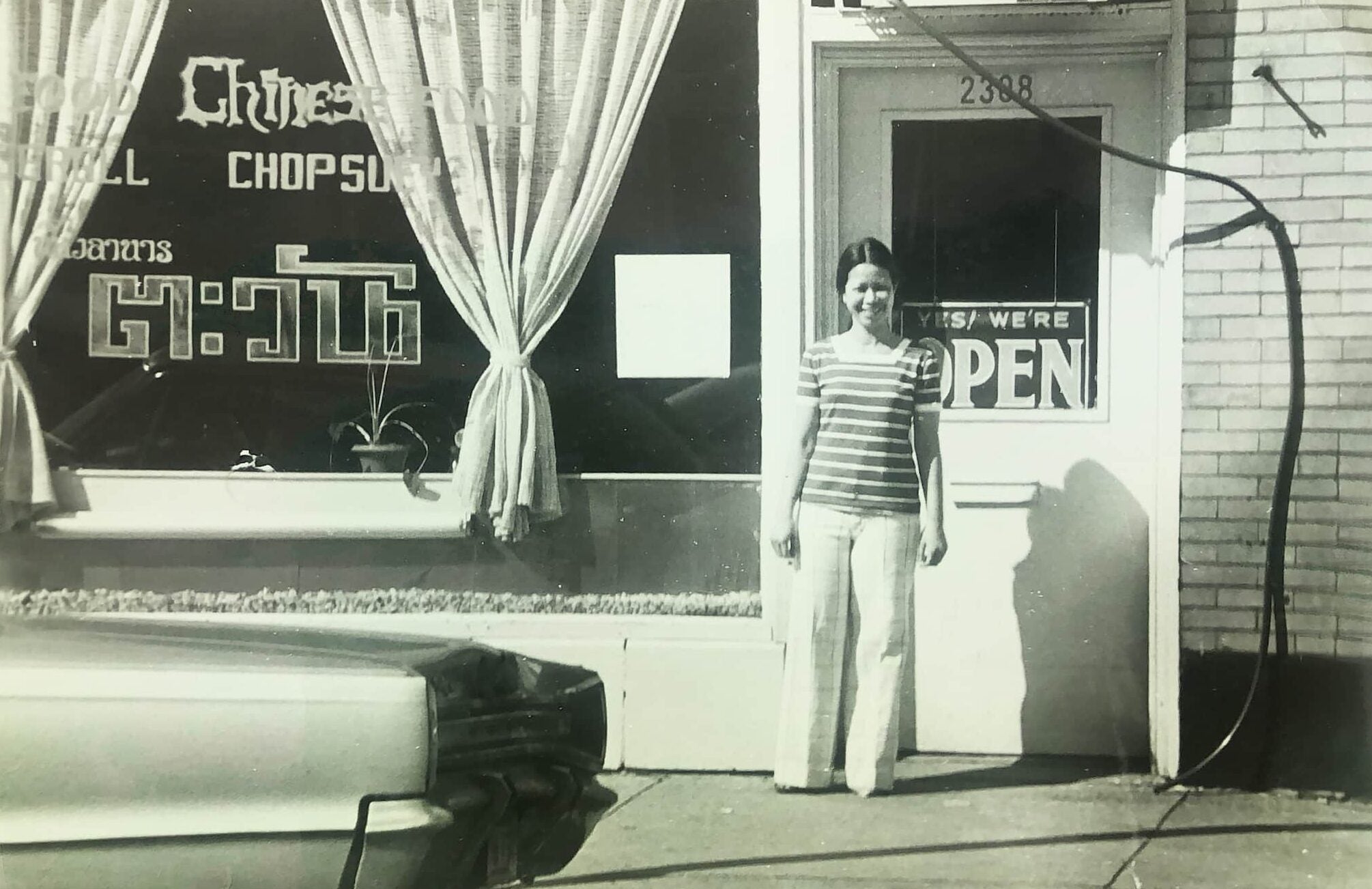 Co-business owner Rushanee Suksod, smiling proudly in a striped shirt, stands by front door of her Thai restaurant Tawan with the open sign displayed.