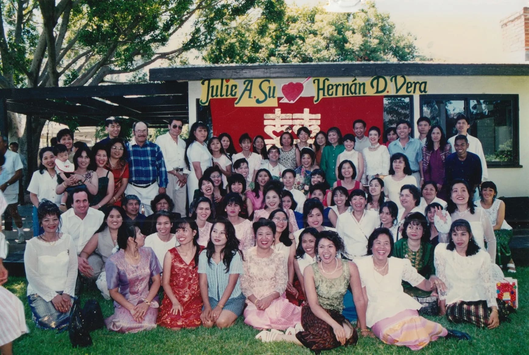 Well-dressed Thai garment workers gather for a large group photo outside in the grass. Some stand, some kneel. A red banner hangs behind them.