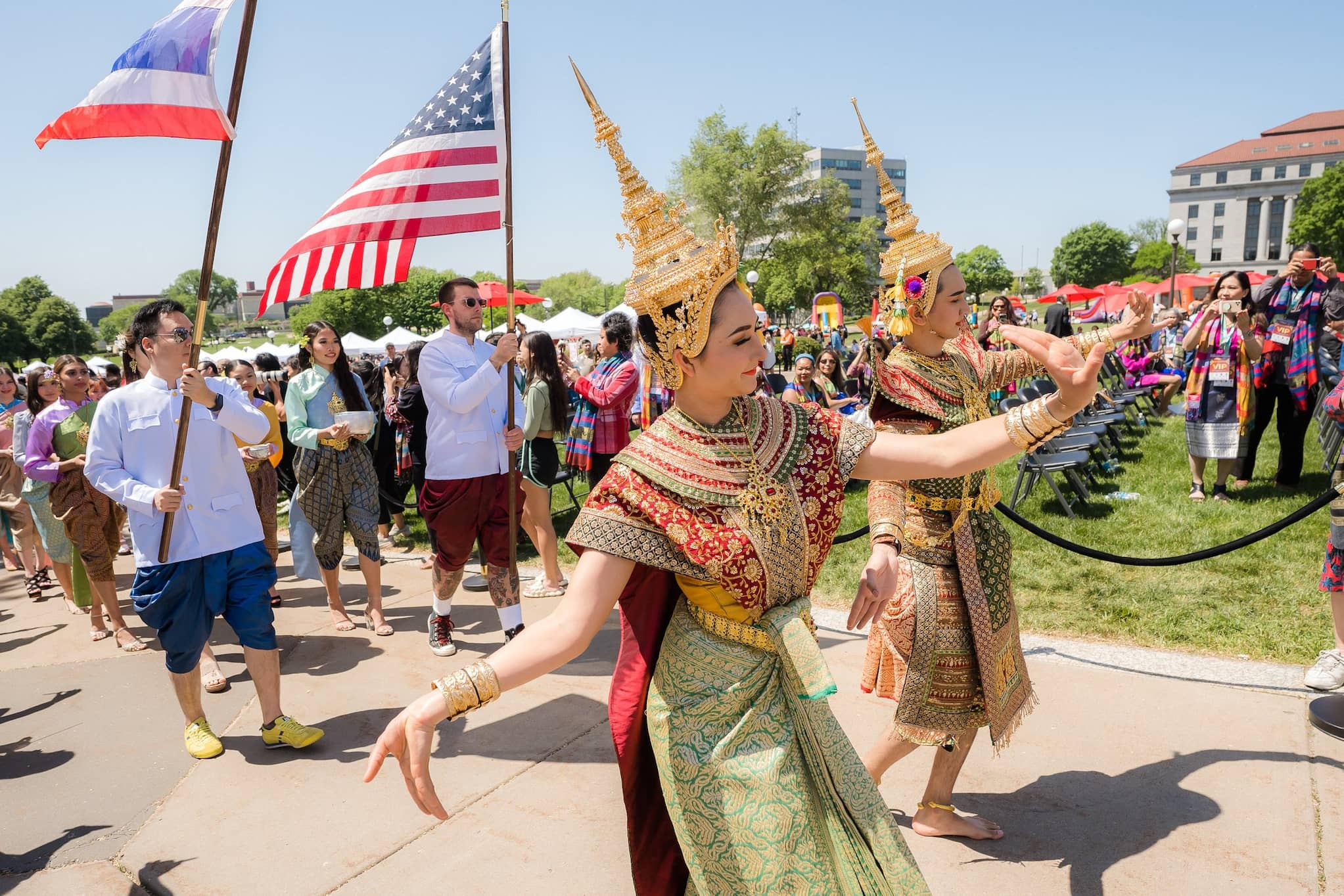 Colorfully dressed Thai dancers with tall, pointed gold headpieces lead outdoor procession, followed by men carrying Thai and US flags.