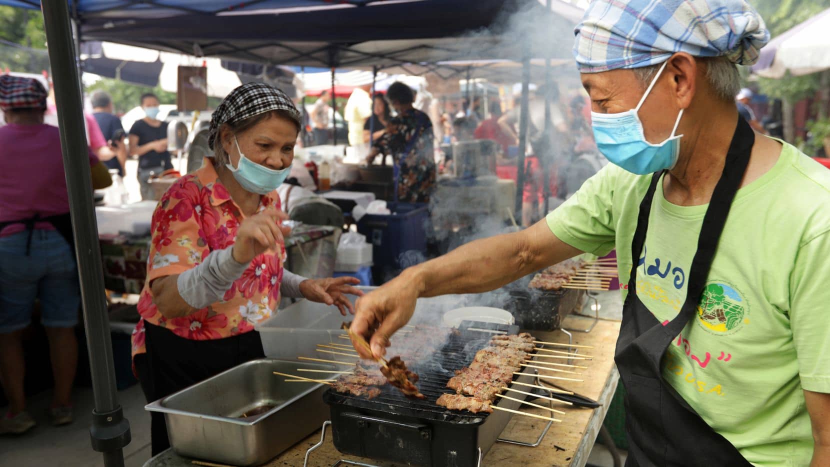 Two food service workers, masked and with hair coverings, prepare skewered meat on grill in outdoor tent. Beside them are more food vendors in tents.
