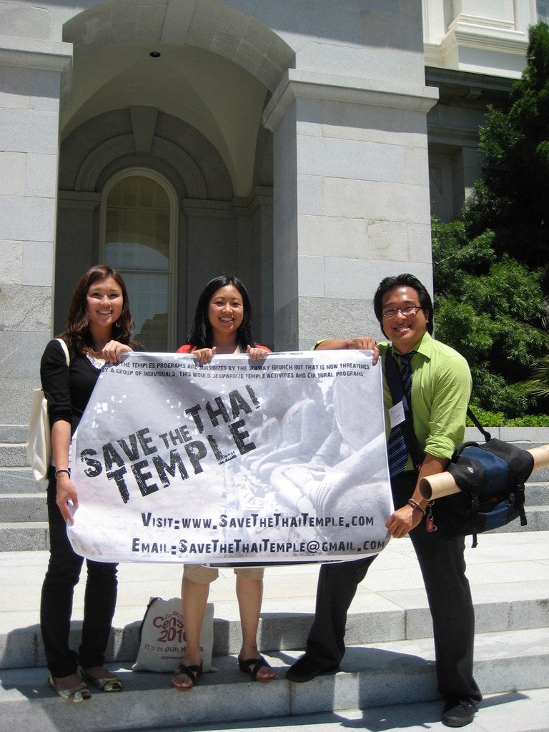 Three people in professional attire stand on steps outside of wat facade. Between them they carry a light gray banner reading "'Save the Thai Temple."
