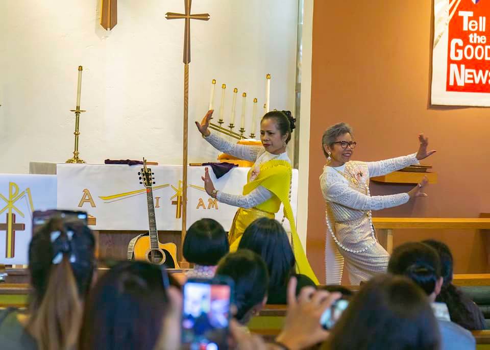 Two women, back to back, perform with arms outstretched on church stage. In foreground, churchgoers in pews watch them dance.