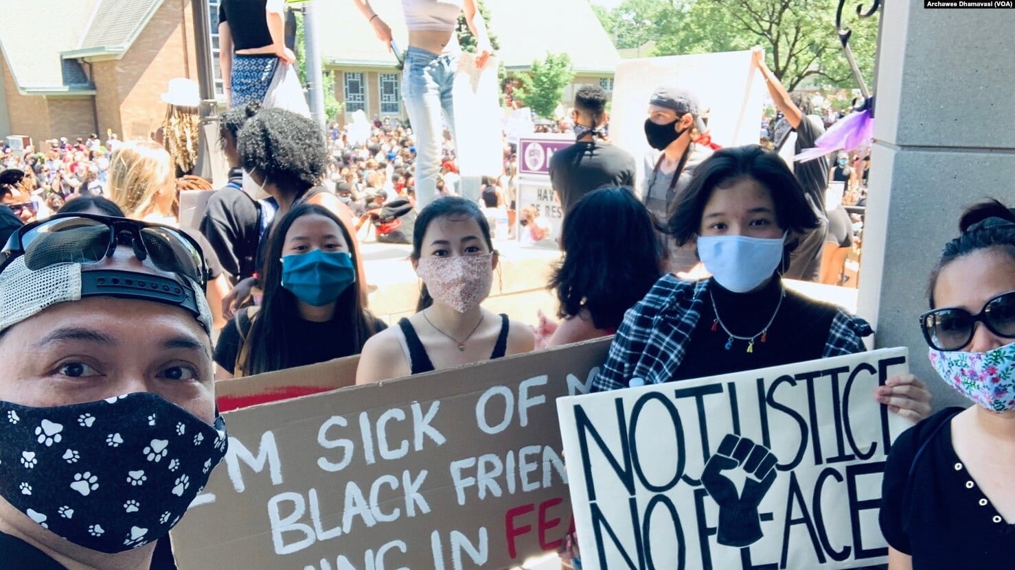 Masked group of Thai Americans holds protest signs during outdoor Black Lives Matter rally. Other rally attendees gather behind them.
