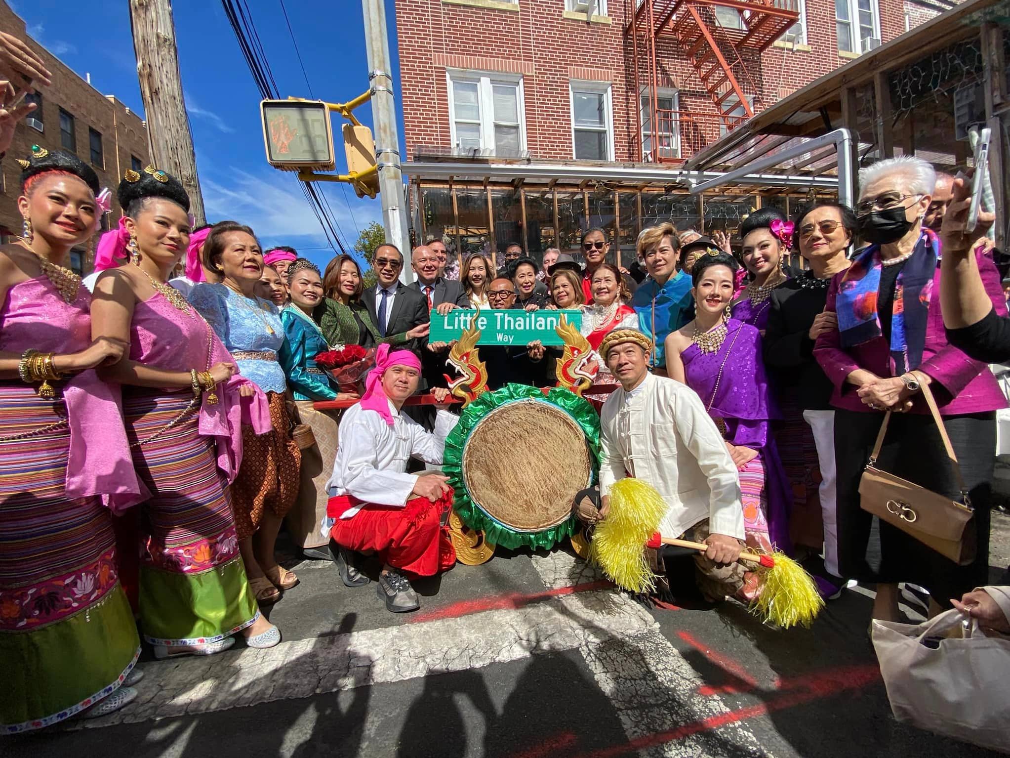 A group of colorfully dressed people crouch and stand near crosswalk. They surround a green street sign reading "Little Thailand Way" in white font.