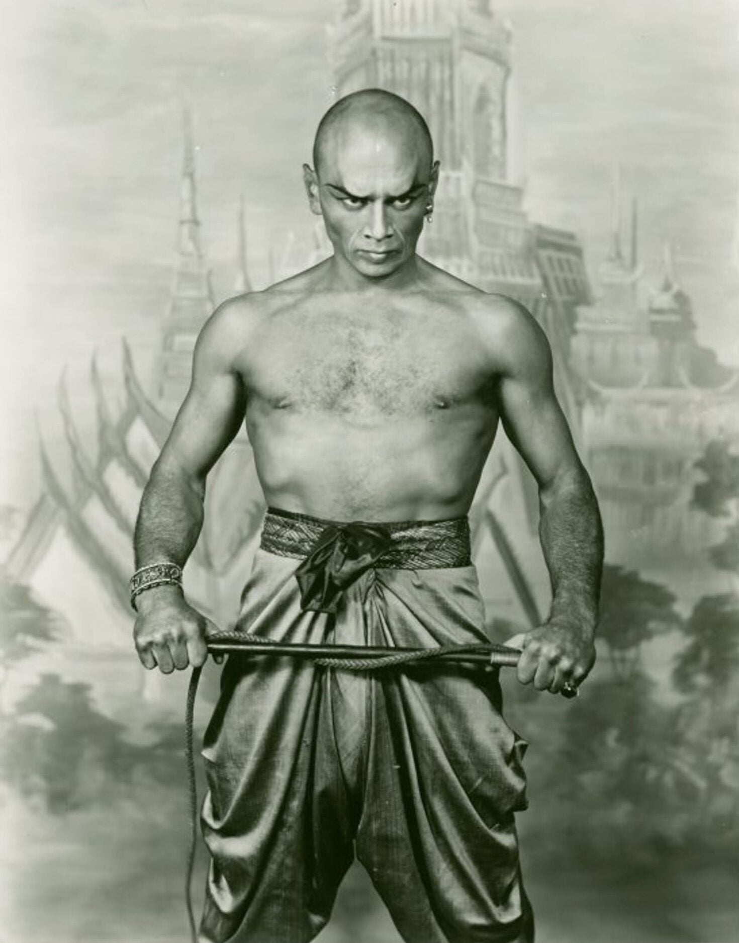 Actor Yul Brynner, a shirtless man with shaved head, poses in character before stage backdrop. Brynner stands with rod clasped between two hands.