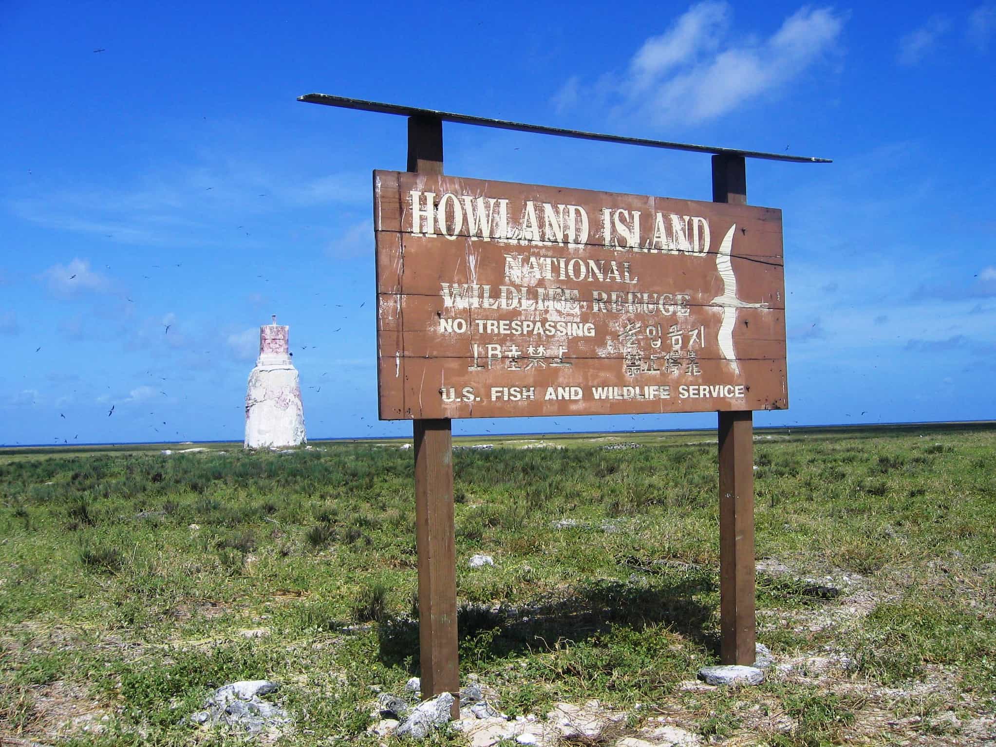 Large, wooden sign with white lettering reads "Howland Island National Wildlife Refuge." Sign is posted outside on flat ground covered in vegetation.