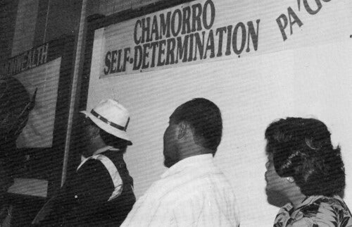 Three people, shown in profile with their faces turned away from the camera, sit beneath sign reading "Chamorro Self-Determination."