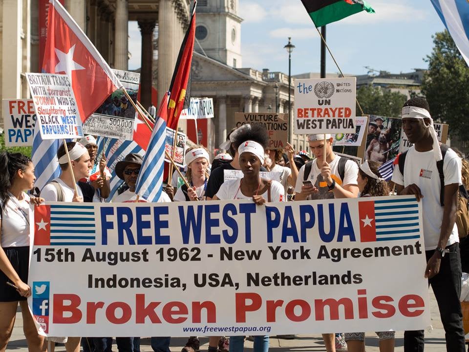 People carrying West Papua flags and signs stand behind large banner reading "Free West Papua" as they gather for outdoor march.