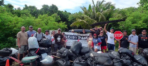 People stand around banner reading "Protect Pagat" as they pose for outdoor group photo. In front of them are piles of large, filled trash bags.