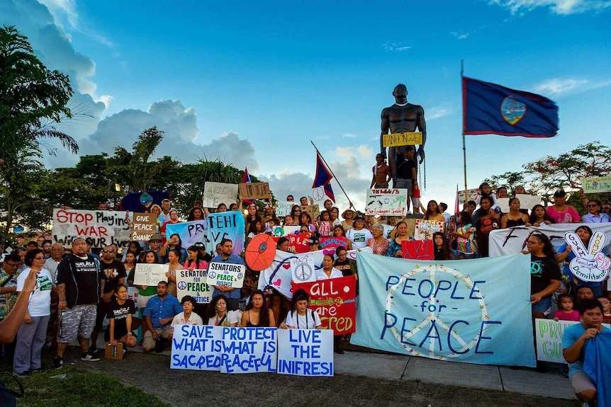 A crowd gathers outside for group photo near statue of standing man. People carry signs calling for peace in Guam, with Guam's flag flying in crowd.