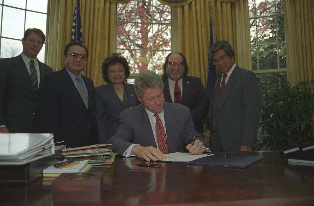 Bill Clinton, seated in the Oval Office, signs the Apology Resolution while Al Gore and the Hawaiʻi congressional delegation surround him.