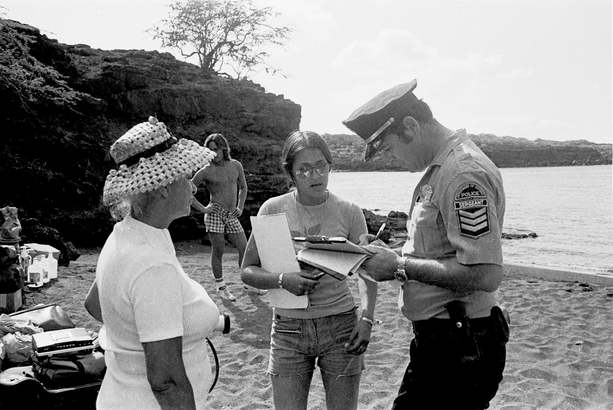 On the shore of Kūheʻeia Bay, a police sergeant writes on a clipboard as two female protestors next to him watch. A shirtless man stands behind them.