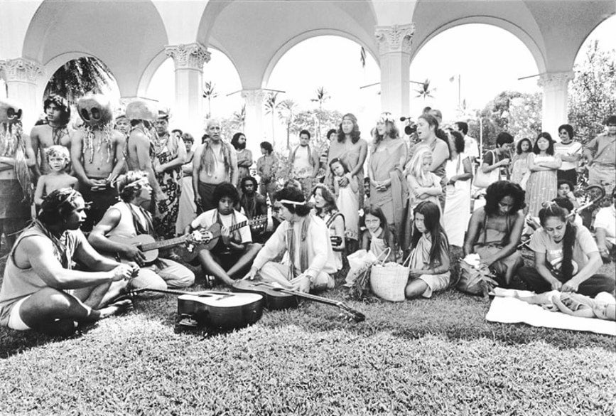 Members and supporters of Protect Kahoʻolawe ʻOhana gather on lawn outside of a federal courthouse with instruments. Some sing and some wear mākini.