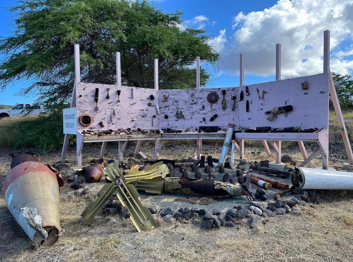 An outdoor display showcases unexploded bombs on a field in Kahoʻolawe. Behind the display is a large tree and a pickup truck.