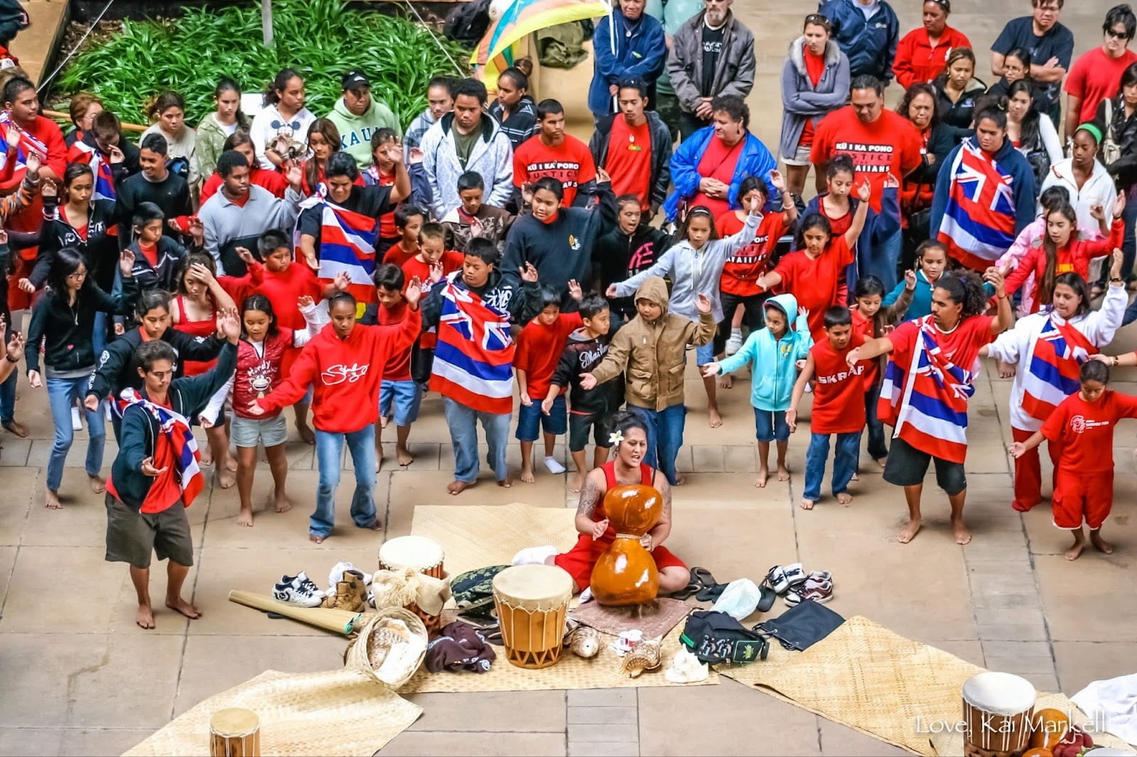Students dressed in Hawaiian flags dance hula kahiko around a kumu hula, a teacher seated on a woven lauhala mat who is singing and playing ipu heke.