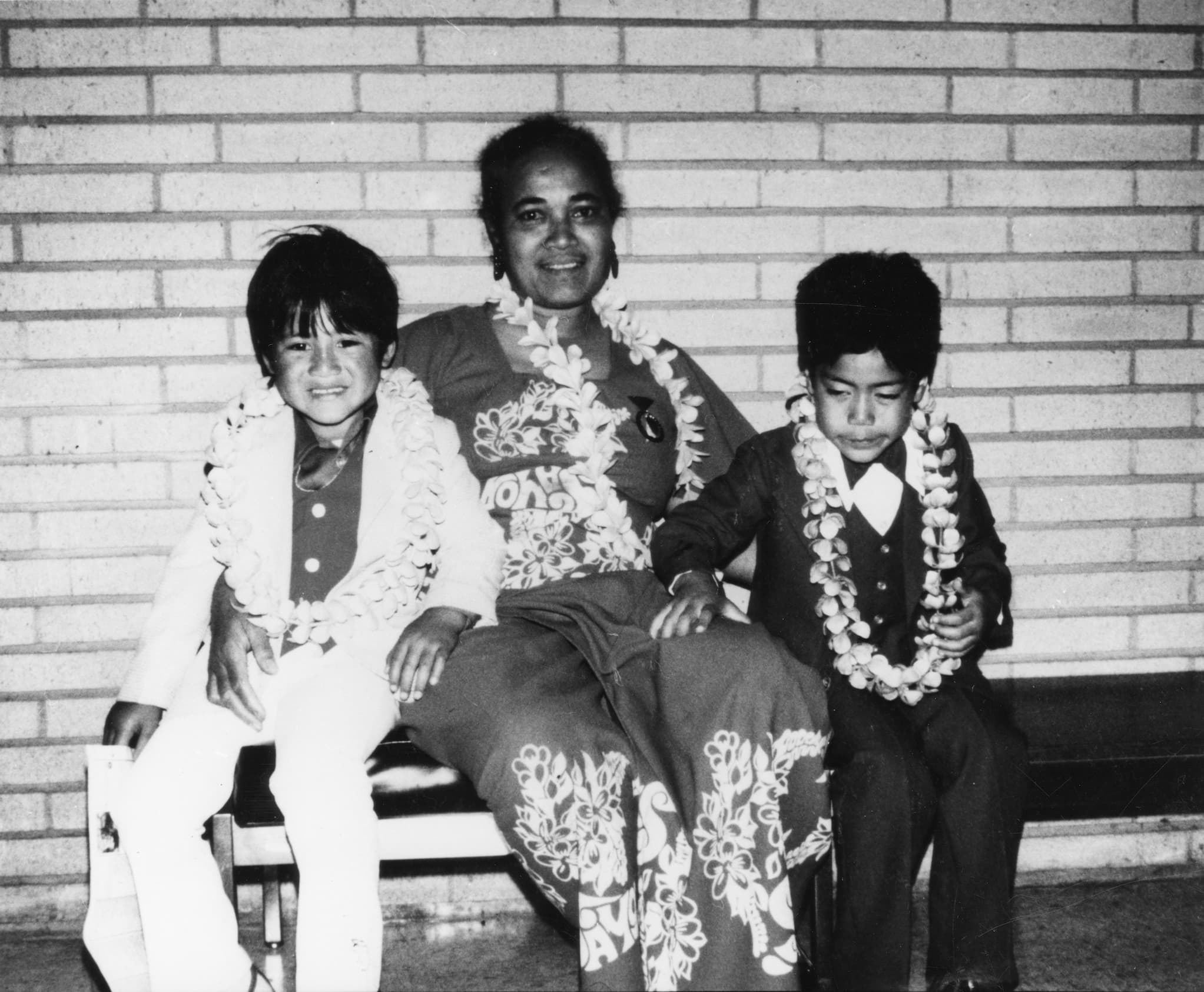 Two Samoan American boys wearing lei and suits sit next to their mother's cousin, wearing a lei and dress, on a bench in front of a brick wall.