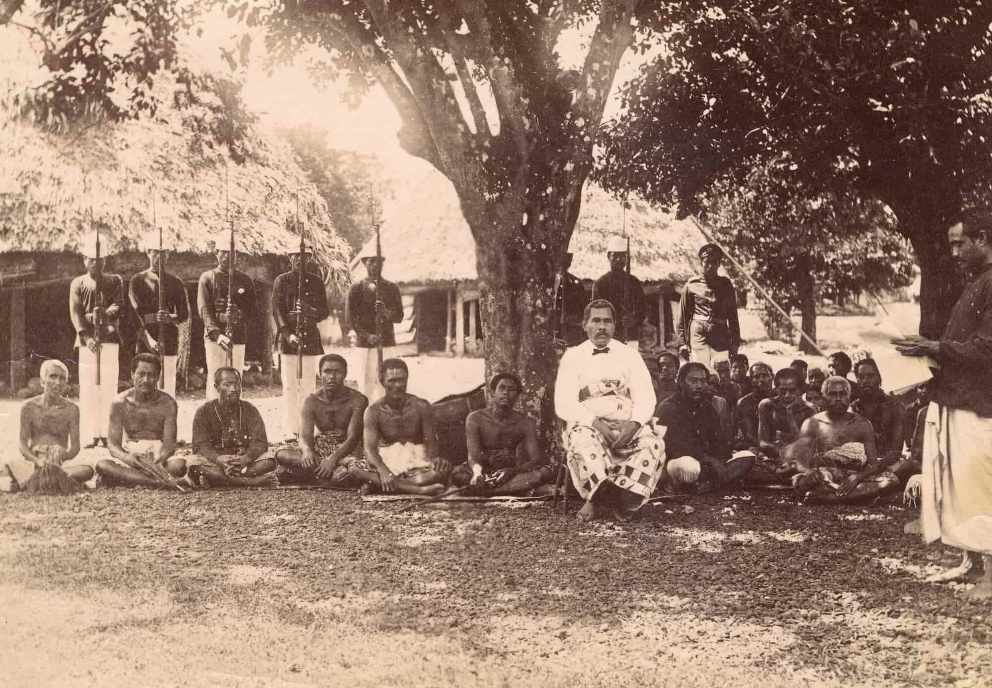 King Malietoa, in white garment, sits on a chair beneath a tree. His followers sit on the ground beside him. Guards with rifles stand behind them.