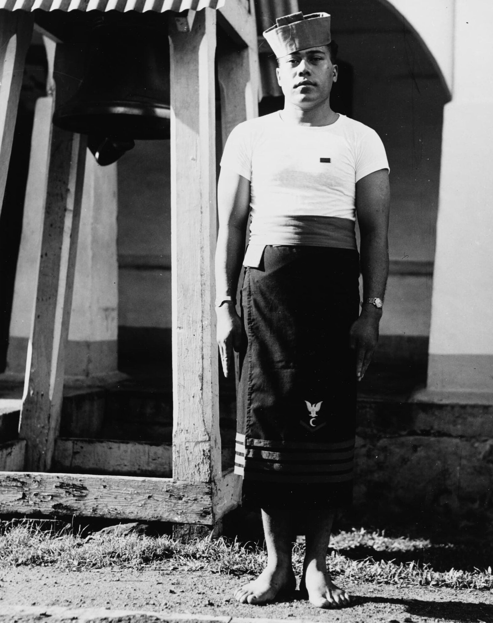A man, dressed in the Fita Fita Guard uniform, stands barefoot at attention next to the wooden bell tower.
