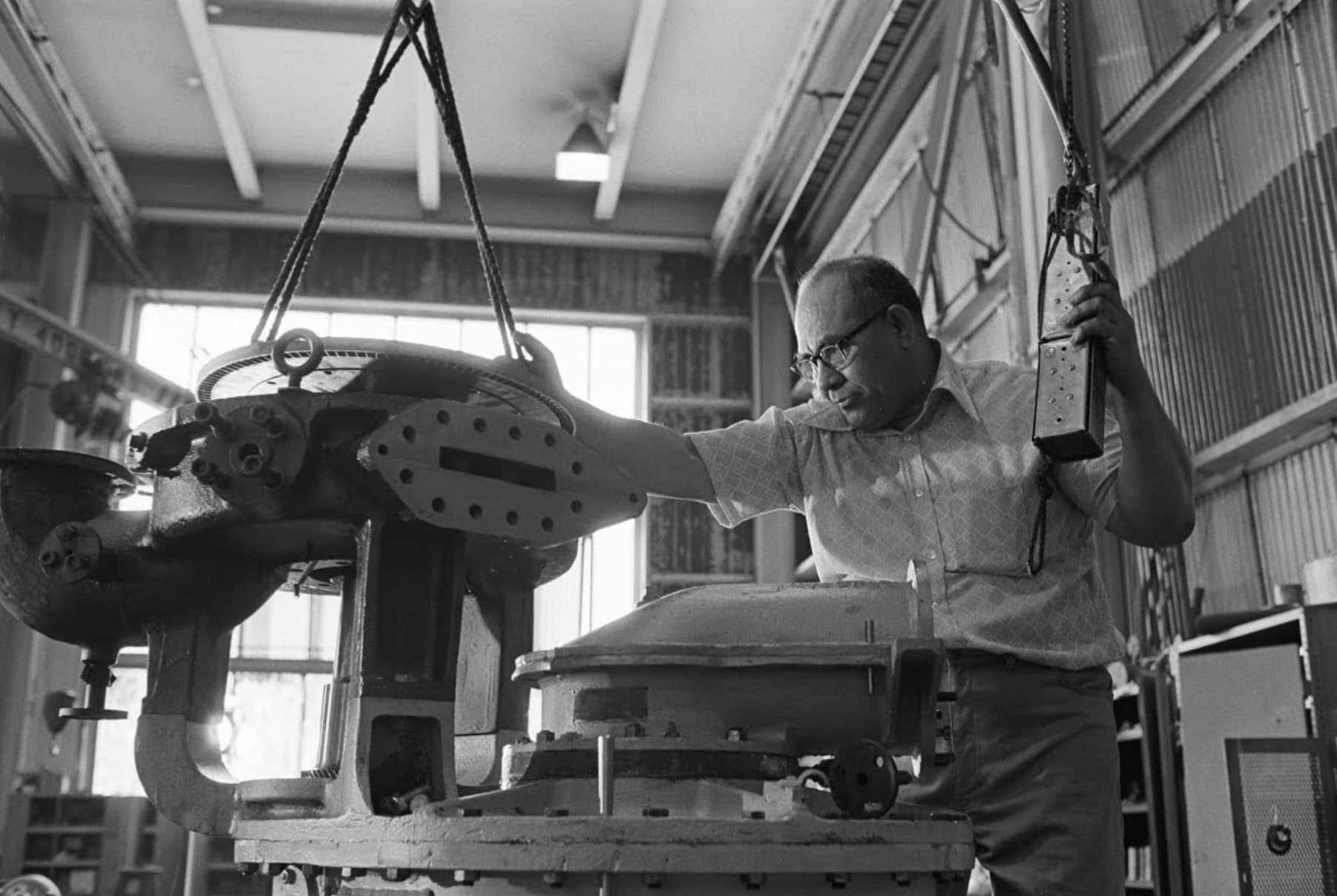 Samoan American shipbuilder Lagofaʻatasi Sialoa, wearing glasses and checkered short sleeve shirt, tinkers on a part of a ship's engine in a workshop.